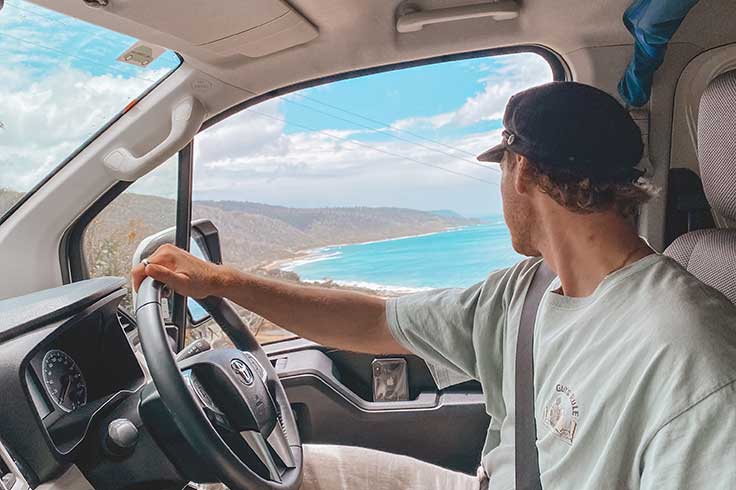 Man holding the steering wheel while looking out of their JUCY Campervan window at the Great Ocean Road views