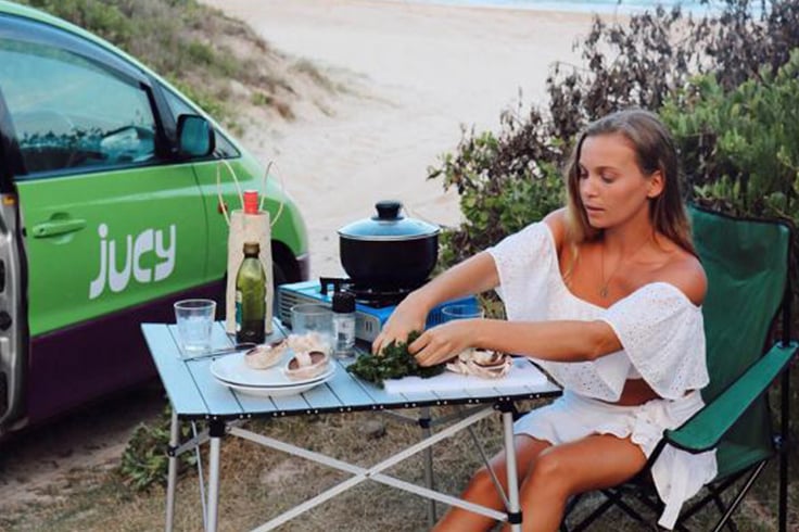 woman eating dinner next to a jucy campervan travelling in summer