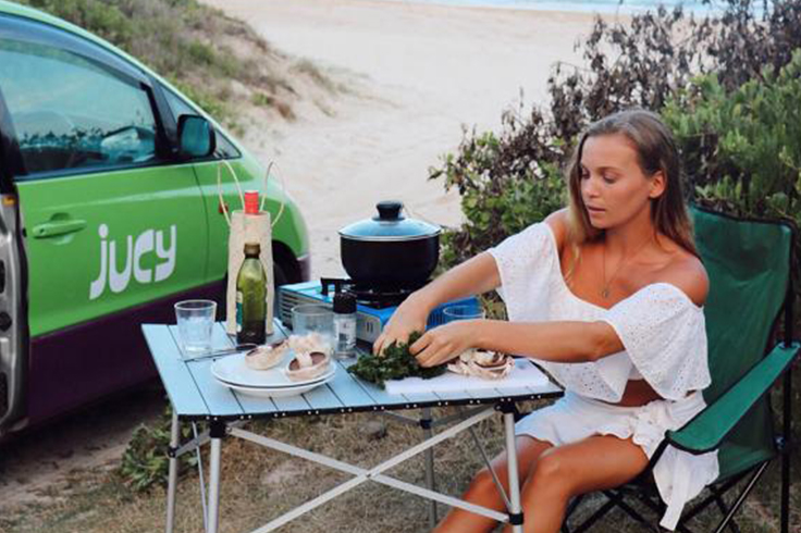 woman eating dinner next to a jucy campervan travelling in summer