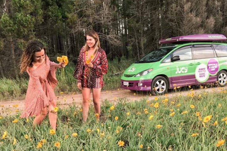 two girls pick spring flowers in a field in australia with a jucy campervan in the background