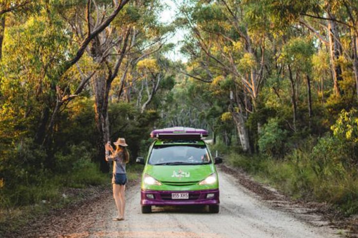 Girl taking photo on a tree lined road with a JUCY campervan