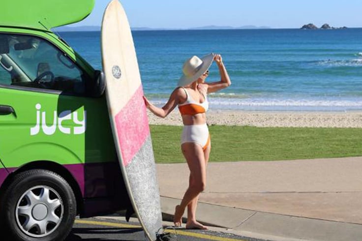 Girl with a surfboard next to a JUCY campervan at a beach
