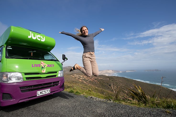 Girl excitedly jumping next to JUCY camper with views of Cape Reinga in the back