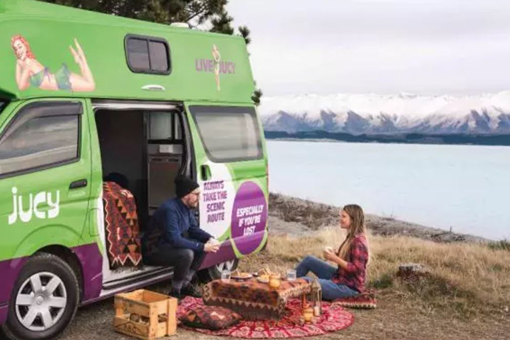 Couple having dinner by a lake with a JUCY campervan