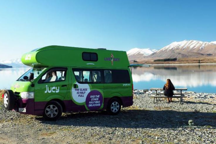 woman sitting at a camping table next to campervan mountains and lake