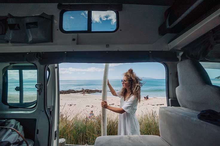 Girl holding surfboard standing outside JUCY Condo campervan