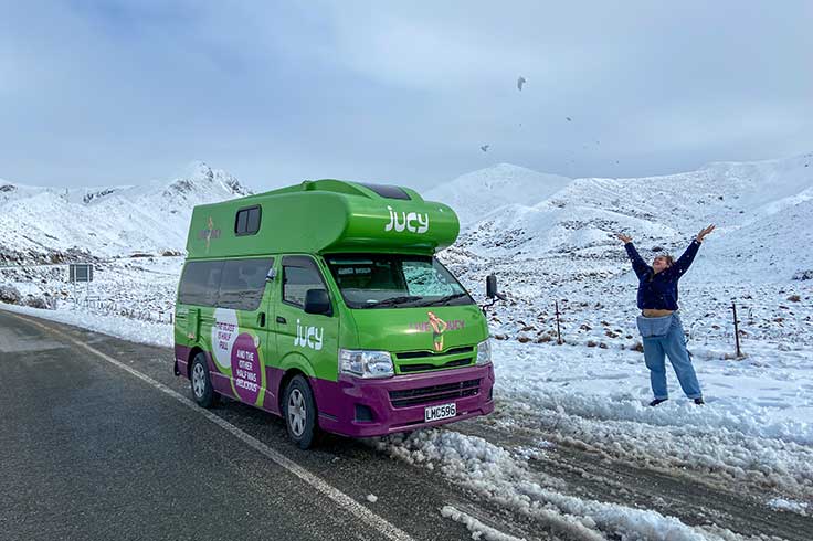 Girl throwing snow beside JUCY Campervan near Lewis Pass