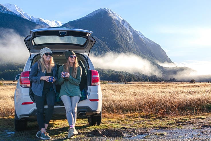 two women drink coffee sitting back of compact suv