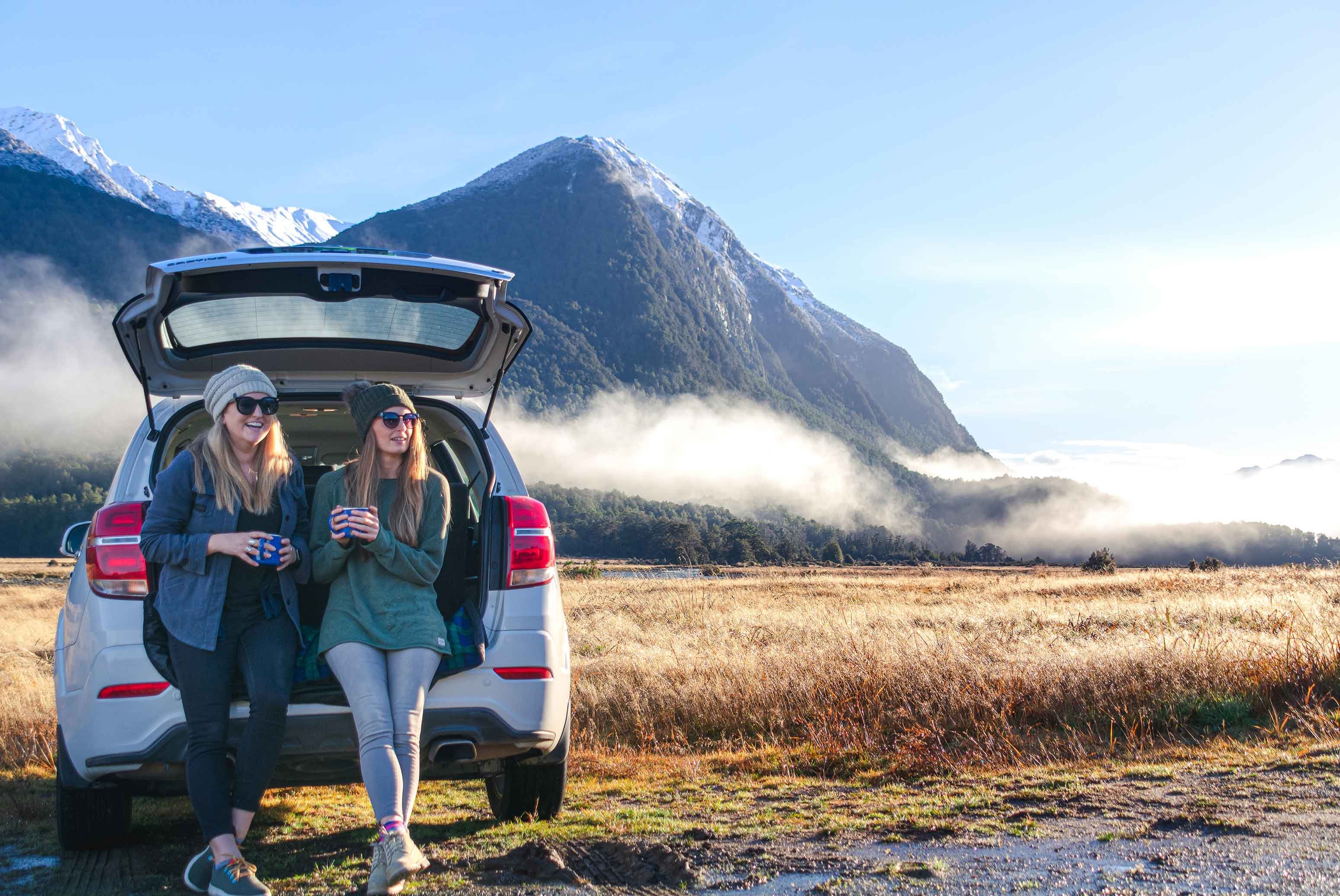 two friends laughing drinking coffee back of compact suv