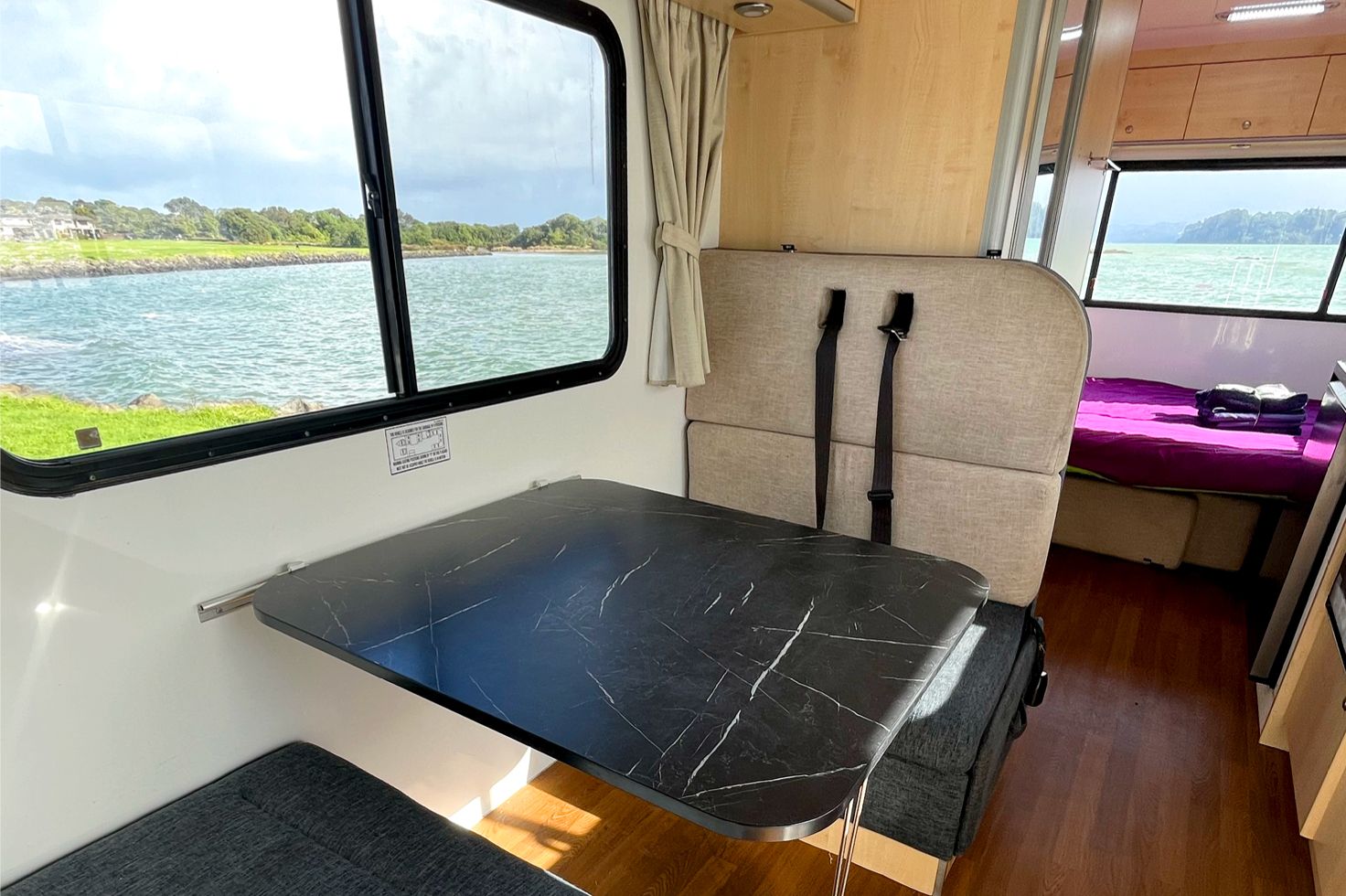 Interior view of a JUCY Big Kahuna motorhome with chairs and table set up next to a window overlooking the sea.